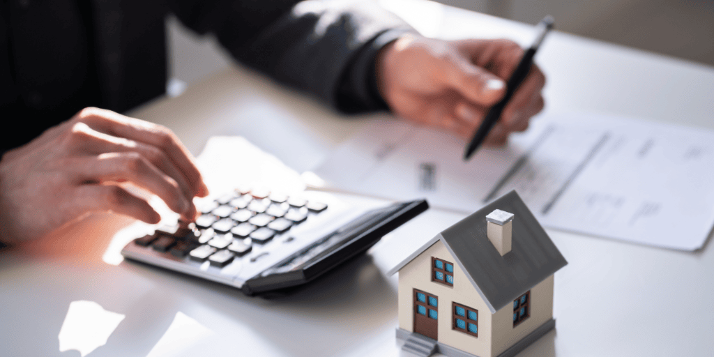 Close-up of hands using a calculator and holding a pen over paperwork, with a small model house on the desk, representing property investment calculations