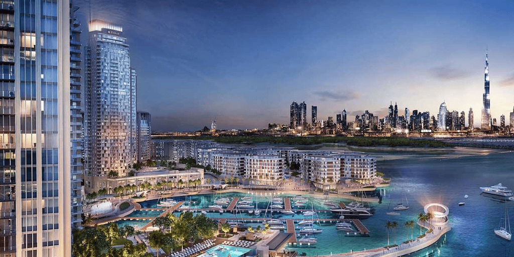 Aerial night view of a waterfront marina development with high-rise residential towers, docked boats, and the Dubai skyline in the background